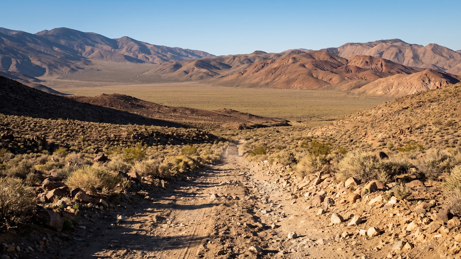 A dusty high-clearance 4x4 truck working up a rough, rocky desert two-track through the open scrub of the Panamint backcountry, banded mountains rising behind