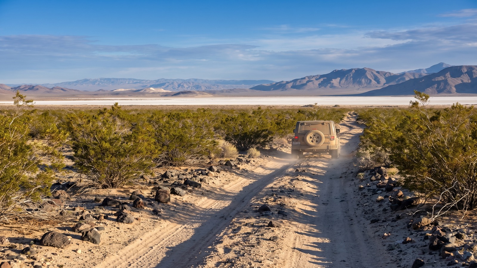 A dusty Jeep heading away down a creosote-lined sandy two-track of the Mojave Road toward a distant dry lakebed and desert mountains