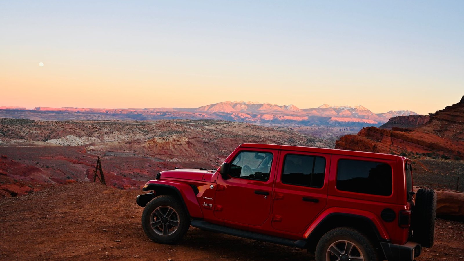 A red Jeep Wrangler on red Moab dirt at dusk with the snow-dusted La Sal Mountains and red-rock formations behind it, the kind of country Pritchett Canyon climbs into