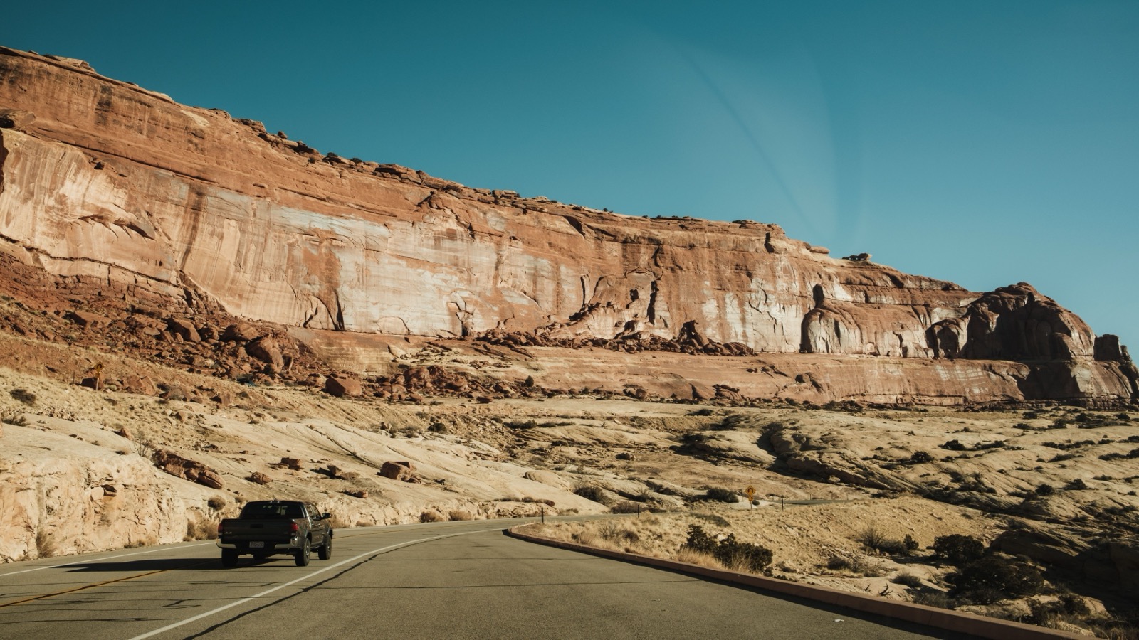 A pickup hugging a towering Moab sandstone wall along a red-rock corridor, typical of the country off Kane Creek Boulevard