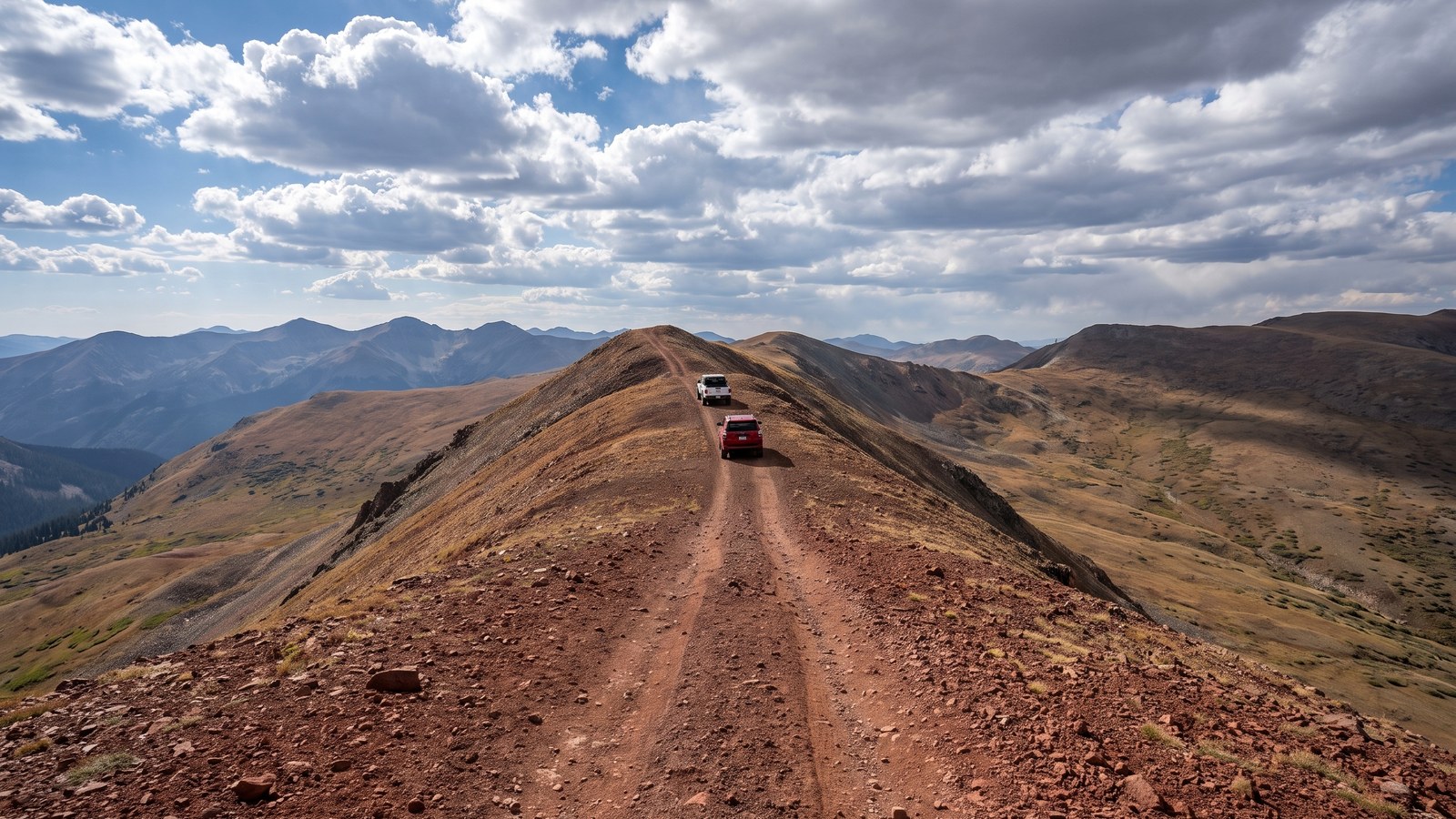 Two 4x4 rigs traversing the narrow red cinder ridgeline of Red Cone above treeline, with a sweeping Continental Divide vista falling away on both sides