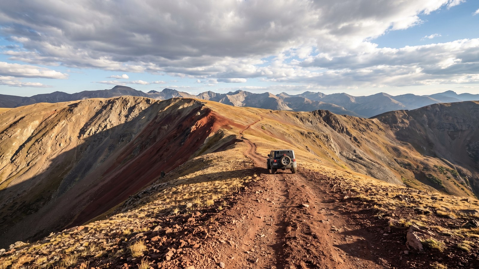 A dark Jeep Wrangler descending the narrow reddish cinder two-track along the exposed Red Cone ridgeline above treeline, with Colorado Front Range peaks stretching to the horizon