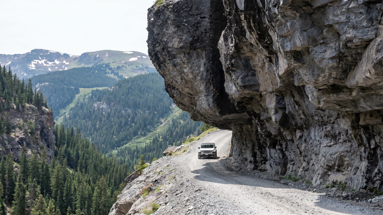 A Jeep passing beneath the massive cantilevered rock overhang on the narrow Camp Bird Road shelf, with the forested Canyon Creek gorge dropping away below and rounded San Juan peaks in the distance
